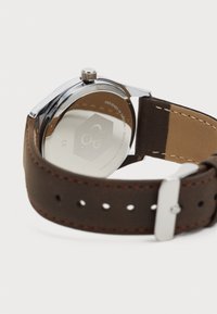 Brown leather strap watch with a silver stainless steel case, visible stitching, and a round, polished back displaying branding and details.