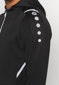 Svart idrottsjacka med dragkedja fram, figurnära design, texturerad tyg och vita accenter med cirkulära mönster på axeln.