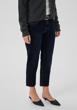 Osoba ubrana w ciemnoniebieskie dżinsy z denimu, czarne, niskie, spiczaste buty, ciemnoszary sweter na zamek i biały top na jednolitym tle.
