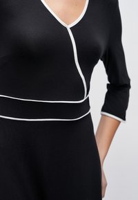 Fekete ruha V-nyakkivágással, háromnegyedes ujjakkal és fehér szegéllyel a varrások mentén. Puha anyagú, testhez simuló szabású, egyszerű sziluett.