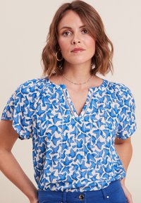 Blauw-witte blouse met korte mouwen en een vogelpatroon, met een V-halslijn en geplooide schouders, gemaakt van lichte stof.