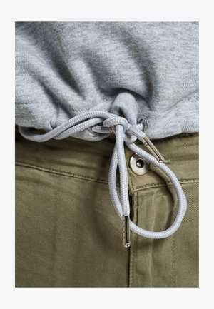 Szürke pulóver anyag, világosszürke húzózsinórral összehúzva, ezüst színű fémalkatrészekkel, olívzöld nadrág felett, amely gombos záródású.