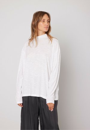 White long-sleeve top with a mock neck, relaxed fit, and lightweight texture. Paired with dark loose-fitting pants for contrast.