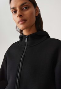 Felpa nera con zip, caratterizzata da un colletto alto, una texture liscia e dettagli delle cuciture sottili. Chiusura con una zip nera sul davanti.