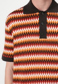 Kortermet polo-shirt med et zigzag-mønster i oransje, rødt, hvitt og svart. Har en mørk krage og treknappslukking.