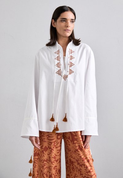 Chemise blanche à manches longues avec des motifs brodés rouille, avec des liens à franges et une coupe ample, associée à un pantalon large imprimé orange.