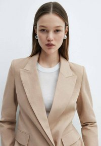 Blazer beige con rever a scendere e due tasche frontali, indossato sopra una maglietta bianca a collo rotondo. Tessuto liscio e design aderente.