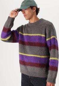 Szürke, bolyhos pulóver széles vízszintes csíkokkal lila, bordó és sárga színben. Bordás szegélye és kerek nyakkivágása van.