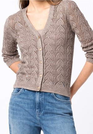 Donna che indossa un cardigan beige lavorato a maglia con chiusura a bottoni e motivi a foglia intricati abbinato a jeans denim azzurri chiari.