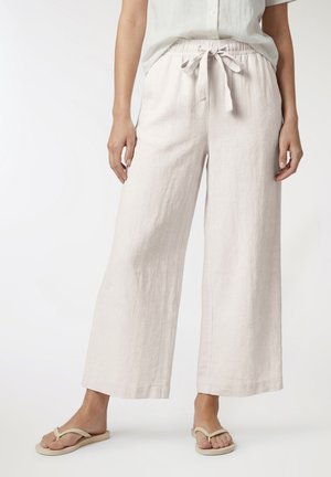 Kobieta w jasnobeżowych lnianych spodniach o szerokich nogawkach z ściągaczem w pasie oraz beżowych japonkach, stojąca na tle białego tła.