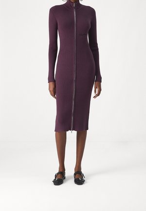 Kvinde iført en tætsiddende, ribstrikket, langærmet, knælængde lilla kjole med lynlås foran og sko med sorte remme og spænder.