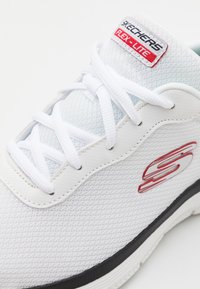 Hvit sportsko med pustende mesh-overdel, flate hvite snører, grå detaljer og en svart såle. "SKECHERS FLEX-LITE"-merke på tungen.
