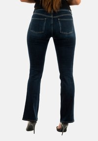 Jeans acampanados de color azul oscuro con una cintura de altura media, cinco bolsillos y detalles de costura sutiles. Combinados con tacones altos negros, mostrando una silueta recta.