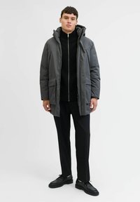 Grå hætteparka med en struktureret overflade, der har en frontlynlås, to store lommer og en sort turtleneck under, parret med mørke bukser.