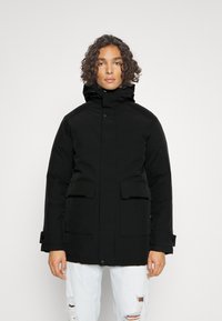 Parka nera con colletto alto, chiusura frontale a bottoni, due grandi tasche e polsini elasticizzati. Tessuto texturizzato, design caldo e silhouette elegante.