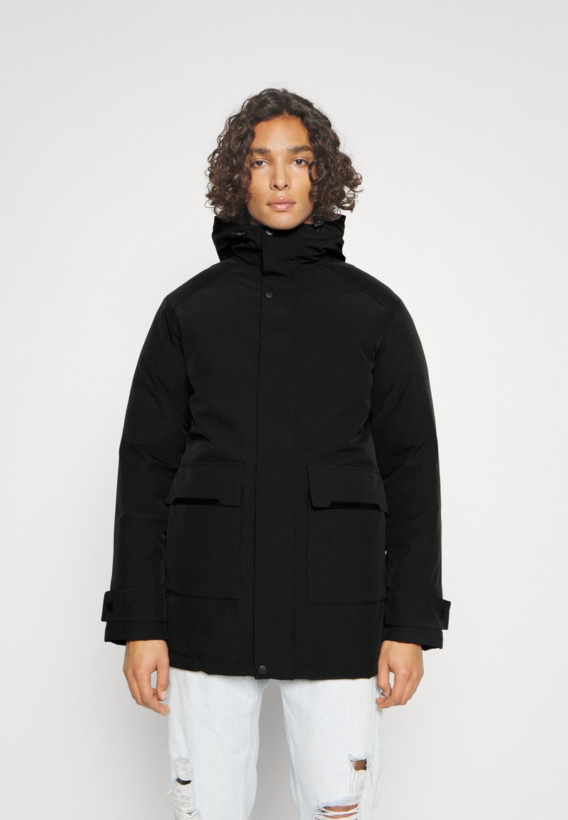 Parka nera con colletto alto, chiusura frontale a bottoni, due grandi tasche e polsini elasticizzati. Tessuto texturizzato, design caldo e silhouette elegante.