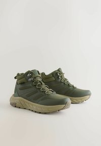 Zielone buty trekkingowe z teksturowaną powierzchnią, wyściełanymi mankietami, wzmocnionymi przelotkami i grubą, wytrzymałą podeszwą zapewniającą przyczepność.