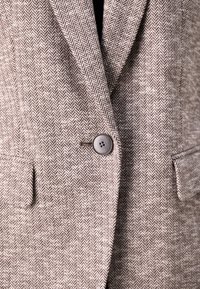 Houndstooth raštas švarkas, naudodamas šviesiai rudos ir tamsiai rudos spalvų paletę, su vienu mygtuko užsegimu ir dviem priekinėmis kišenėmis. tekstūruotas audinys.