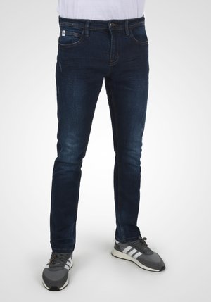 Jean en denim foncé avec une coupe ajustée, comprenant cinq poches, un léger délavage et une fermeture classique à bouton et fermeture éclair. Porté avec des baskets grises.