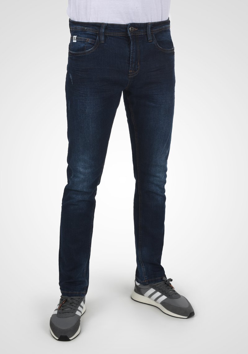 Mørke denimjeans med slim fit, som har fem lommer, subtil falming og en klassisk knap- og lynlåslukning. Båret med grå sneakers.