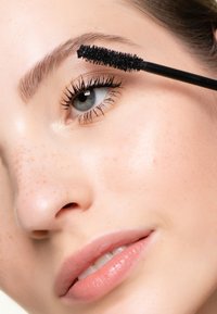 Spazzola di mascara nera che applica prodotto su lunghe ciglia scure. Pelle chiara con lentiggini, colore delle labbra naturale e sfondo neutro.