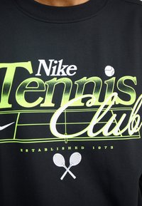 Melns džemperis ar izteiksmīgu baltu un neona zaļu "Nike Tennis Club" uzrakstu, tenisa bumbiņas grafiku un krustotu raketi. Izveidots 1972.