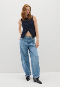 Top senza maniche blu navy con dettagli a bottoni e design corto, abbinato a jeans azzurri larghi. La modella indossa stivali neri con tacco.