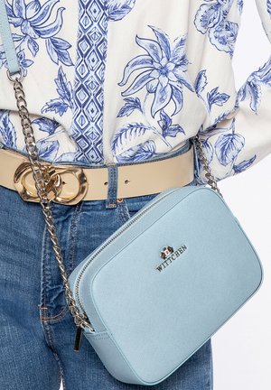 Světlomodrá kabelka přes rameno s texturovaným povrchem, stříbrným řetízkovým popruhem a jemným logem. Kombinována s květinovou blůzou a béžovým pásem.