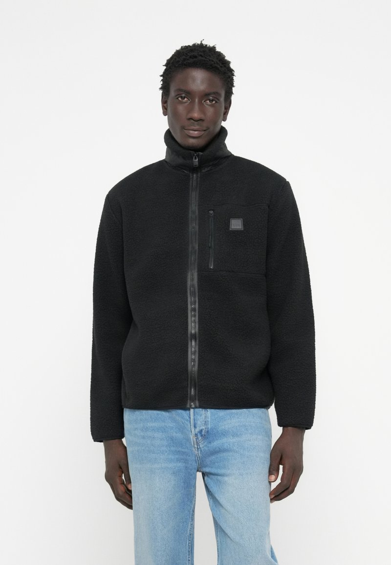 Black fleece jacket with high collar, zip closure, and a chest pocket. Soft texture and relaxed fit. Paired with light blue denim jeans.