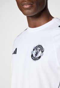 Bijela atletska majica sa crnim Adidasovima logotipom na ramenima i izvezenim grbom Manchester Uniteda na prsima.