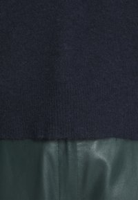 Zbliżenie na ciemnoniebieską dzianinę umieszczoną nad gładkim, ciemnozielonym materiałem skórzanym z widocznym pionowym przeszyciem.