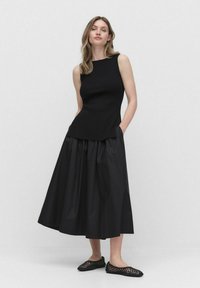 Czarny bezrękawnik o ryflowanej teksturze, w zestawie z długą czarną plisowaną spódnicą. Stylizacja dopełniają płaskie czarne plecione buty.