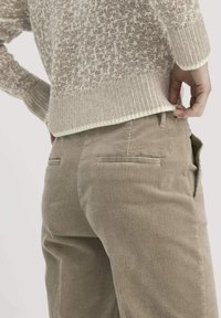 Persoon in beige corduroy broek en een gestructureerde gebreide trui, die met één hand de onderkant van de trui aanpast.