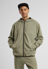 Oliwkowy hoodie zapinany na zamek, wykonany z miękkiego materiału, z kapturem, przednimi kieszeniami i czarnym akcentem zamka błyskawicznego. Posiada teksturowaną podszewkę.