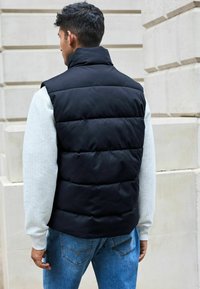 Zwarte gewatteerde gilet met hoge kraag, gecombineerd met een lichtgrijze gestructureerde trui en blauwe jeans. Van achteren gezien tegen een lichte muur.