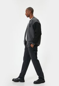Chaqueta varsity negra y gris con la letra "W", combinada con pantalones negros y botas negras gruesas. Tejido suave con puños acanalados.