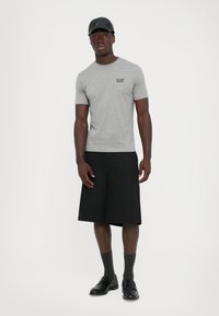 Mann står iført en grå EA7 t-skjorte, svarte vide shorts, grå sokker, svarte loafers og en svart caps mot en ensfarget hvit bakgrunn.