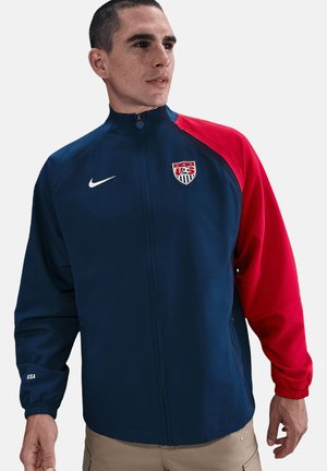Giacca Nike navy e rossa con colletto alto, chiusura a zip e polsini elasticizzati. Presenta il logo USA e della nazionale di calcio degli Stati Uniti sul petto. Materiale leggero.