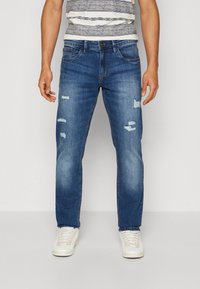 Jeans in denim blu con dettagli strappati, caratterizzati da una gamba dritta, cinque tasche e chiusura con zip e bottone. Indossati con sneakers bianche.