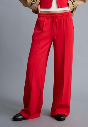 Személy élénk piros, széles szárú nadrágban, amelynek elasztikus derékpántja és húzózsinórjai vannak, fekete cipőben, bézs kabátban és piros, rövid felsőben, szürke háttér előtt.