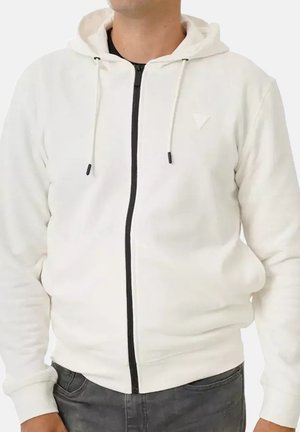 Homme portant un sweat à capuche blanc avec fermeture éclair noire et cordons, associé à un jean gris foncé, sur un fond uni.