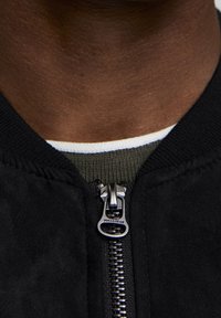 Blouson aviateur en daim noir avec col côtelé, doté d'une fermeture éclair argentée. Chemise à rayures vertes et blanches superposée visible en dessous.