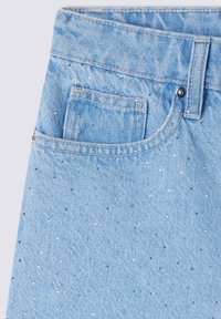 Tessuto di denim azzurro chiaro con un motivo maculato di borchie argentate. Presenta una tasca frontale e una chiusura con bottone in metallo. Texture strappata vicino alla vita.