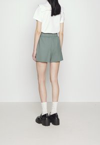 Top bianco a maniche corte con dettagli in pizzo sulle spalle, abbinato a pantaloni corti verdi. Il modello indossa scarpe con platform nere e calze bianche da tennis.