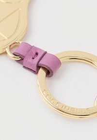 Χρυσό μπρελόκ συνδεδεμένο με ροζ δερμάτινη λωρίδα με ορατές ραφές, χαραγμένο με τη λέξη "COCCINELLE" στον μεταλλικό κρίκο.