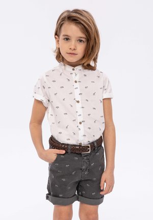 Camicia bianca a maniche corte con stampa animale grigia e bottoni marroni, abbinata a pantaloni corti antracite con orlo arrotolato e cintura intrecciata marrone.