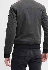 Un uomo indossa una giacca bomber grigio scuro e jeans neri, visto di spalle, mentre sta in piedi contro uno sfondo chiaro e uniforme.