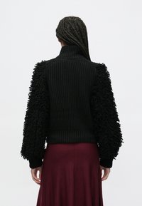 Černý pletený svetr s texturovanými, chlupatými rukávy, vysokým límcem a žeberným tělem. Nošen s bordó sukní, která ukazuje vrstvený styl.