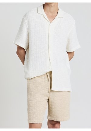 Homme portant une chemise crème texturée à manches courtes boutonnée et un short en tricot beige avec cordon de serrage sur fond uni.