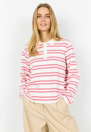 Žena oblečená v bielej polokošeli s ružovými vodorovnými pruhmi a béžovými nohavicami, stojaca s rukami v kapse.